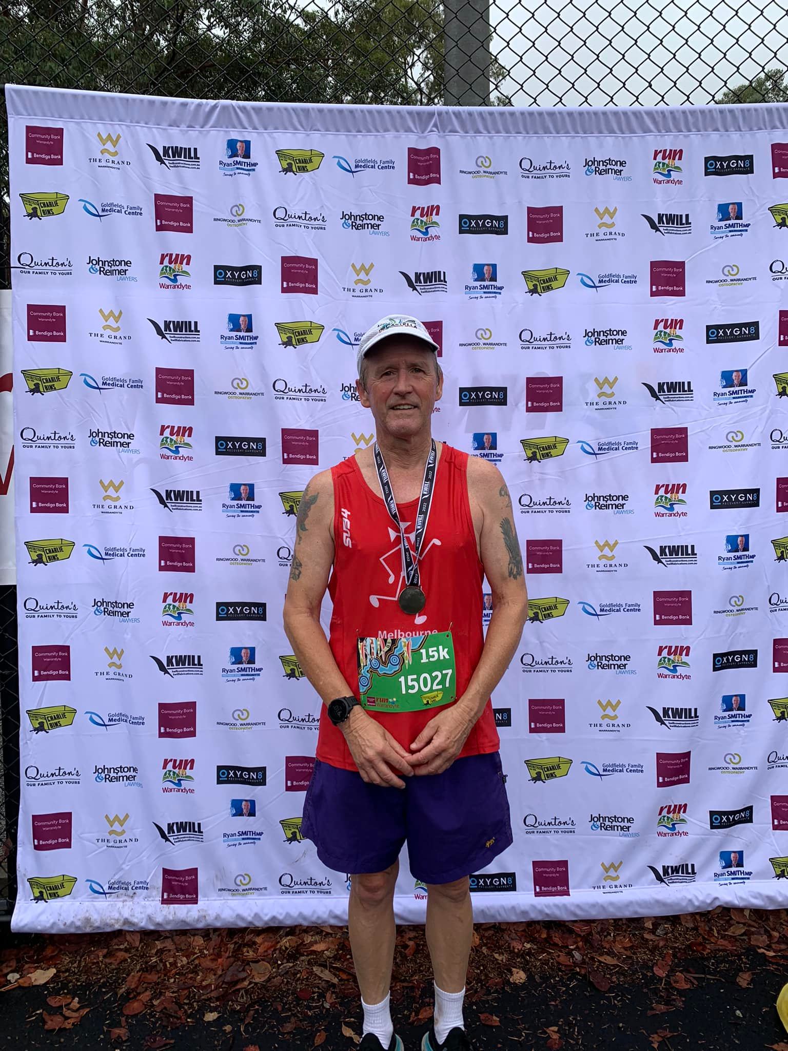 A runner in red singlet with a medal and a racing bib in front of a sign with the names of sponsors